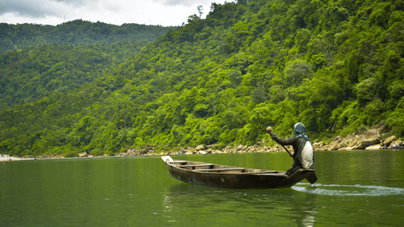 Lone boatman in a wooden canoe rowing across a green river with dense forested hills rising behind under a cloudy Meghalaya sky.
