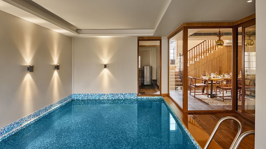 Indoor swimming pool with blue mosaic tiles, modern wall lighting, and a metal pool ladder. A glass partition separates the pool area from a cosy dining room with wooden furniture and warm lighting - Voyage The Solitaire Bliss.