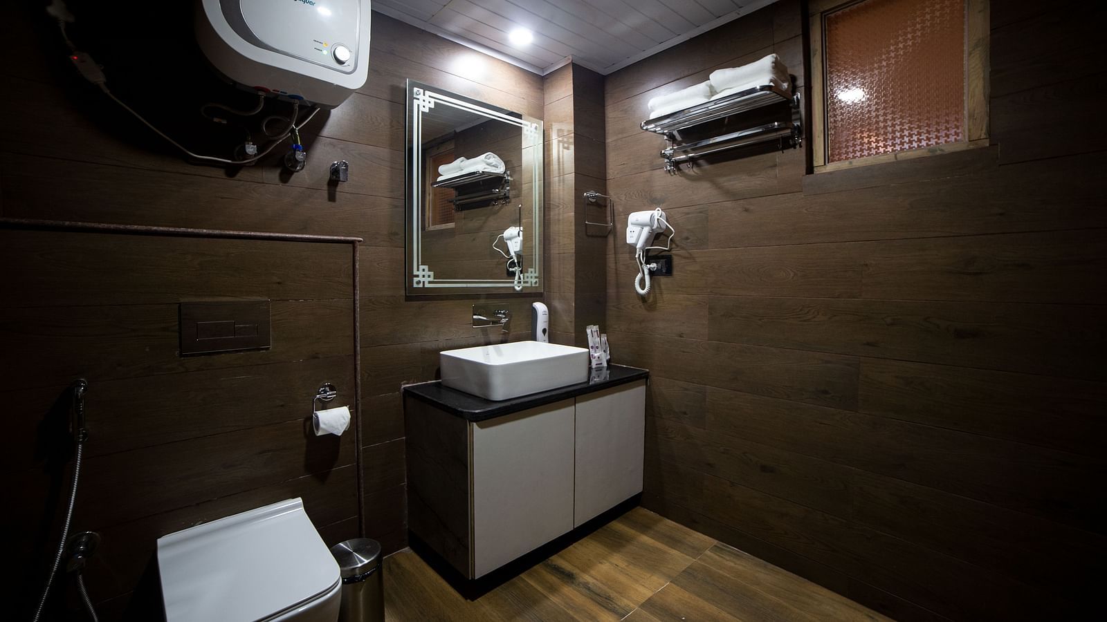 Contemporary hotel bathroom with dark wood-style walls, a wall-mounted toilet, a modern vanity with a square sink and large mirror, and amenities including a hairdryer, towel rack, and water heater - Yashshree Lachen.