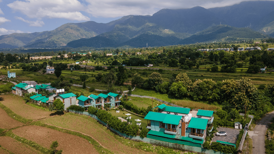 Aerial view of Corbett Nirvana Resort, Uttarakhand featuring cottage style buildings in clusters surrounded by farmlands, trees, and a backdrop of green hill.