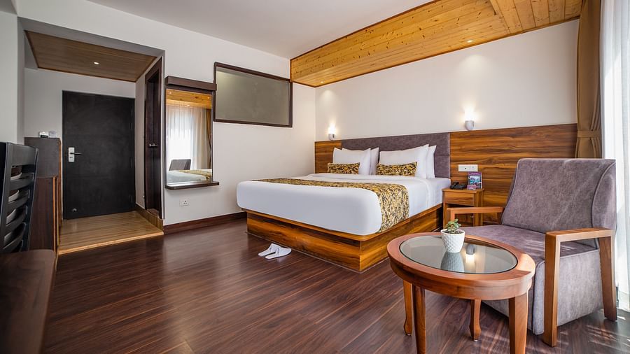 A  deluxe room in Sumitel Hotel with double bed, a lone chair in the corner, a circular table in front of the chair, a TV, and a mirror.