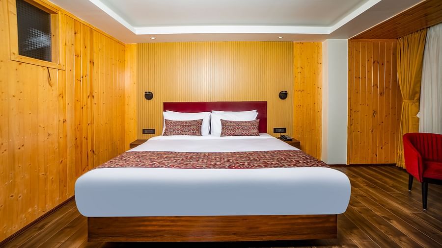 Neat and cosy hotel room featuring a large double bed with white and maroon bedding, wooden panel walls, warm lighting, and polished wooden flooring, creating a welcoming atmosphere - Yashshree Lachen.