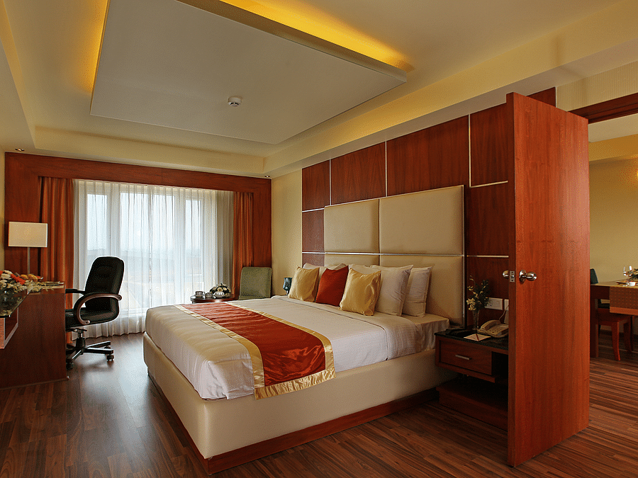 A spacious hotel suite with a plush king-size bed, wooden flooring, and warm lighting, featuring a work desk and a large window. - La Classic, Hosur Road
