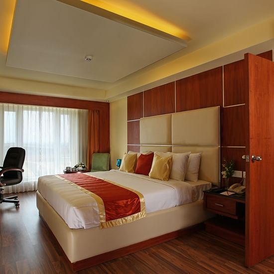 A spacious hotel suite with a plush king-size bed, wooden flooring, and warm lighting, featuring a work desk and a large window. - La Classic, Hosur Road