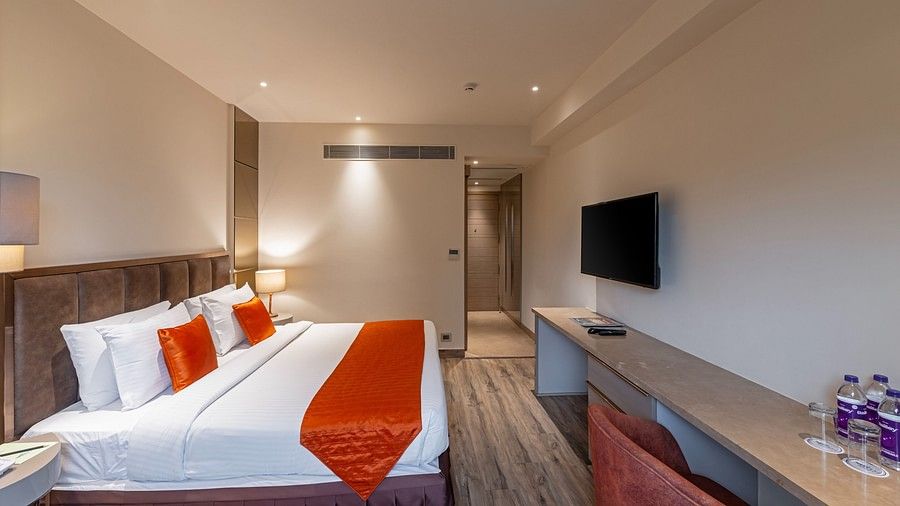A bright bedroom featuring white ceiling lights and lamps, with natural light filtering in through the window. The bed is positioned against one wall, facing a TV mounted on the wall above a desk and matching chairs with water bottles on top.