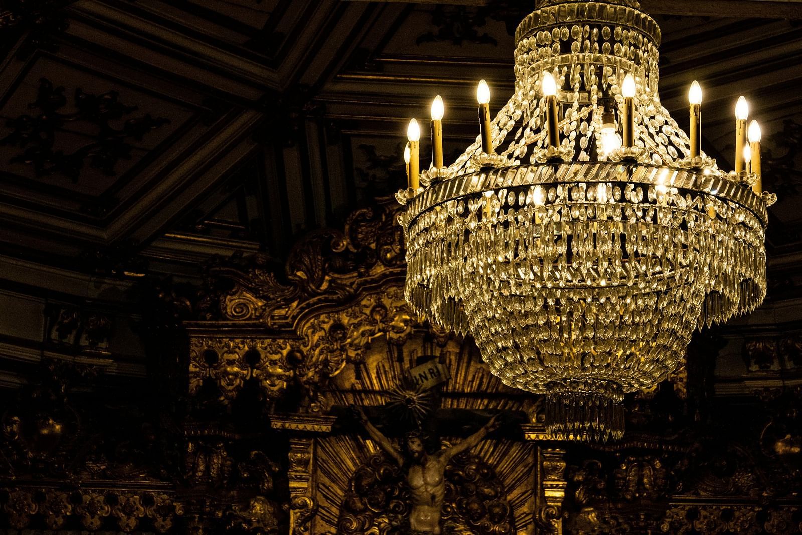 A grand, multi-tiered chandelier with numerous lights hangs in a dimly lit, ornate space.
