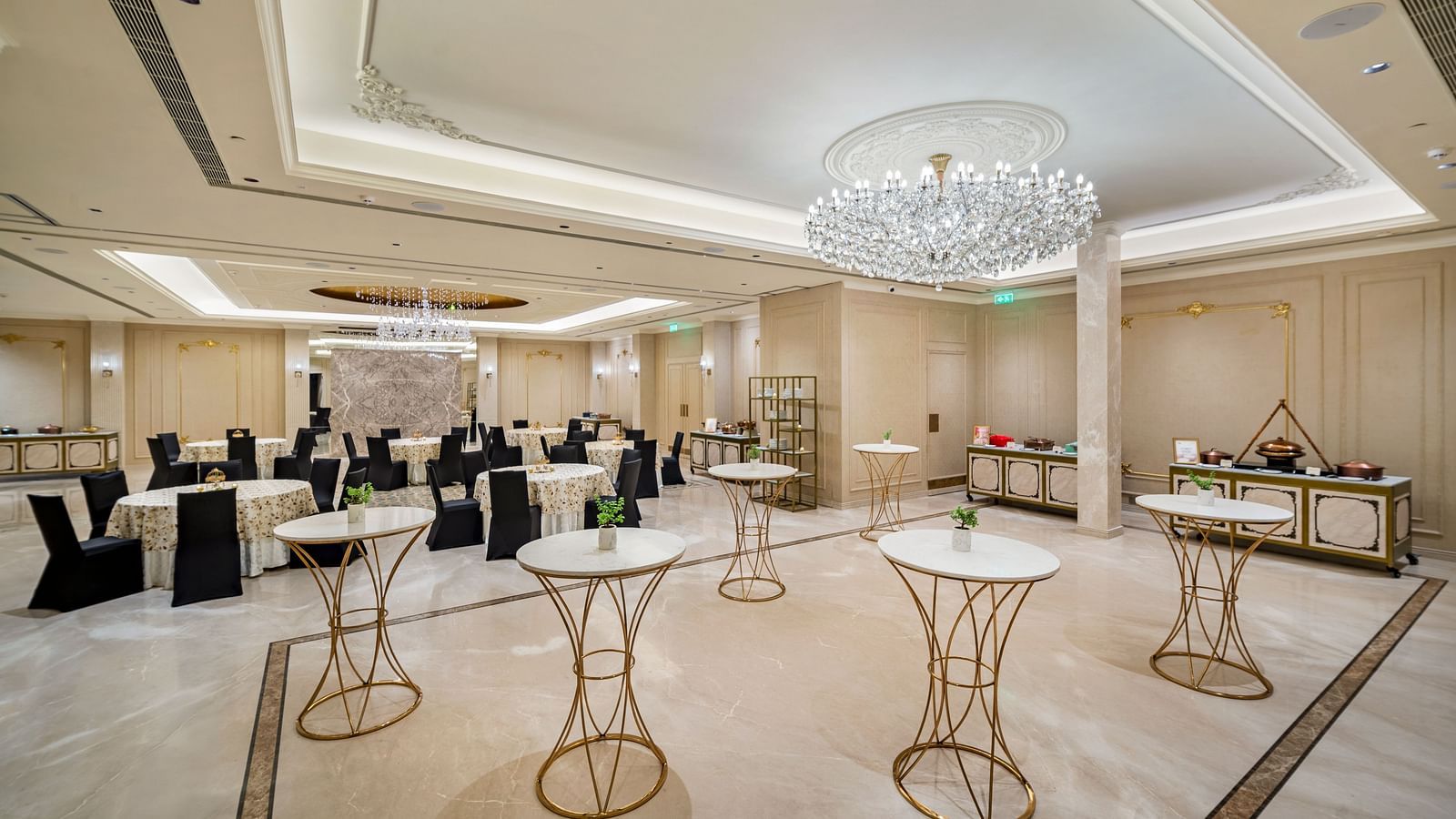An elegant pre-function or cocktail area at Grand Chennai by GRT Hotels featuring numerous white and black high-top tables, perfect for networking or informal gatherings. A grand, modern chandelier illuminates the spacious room with a soft light.