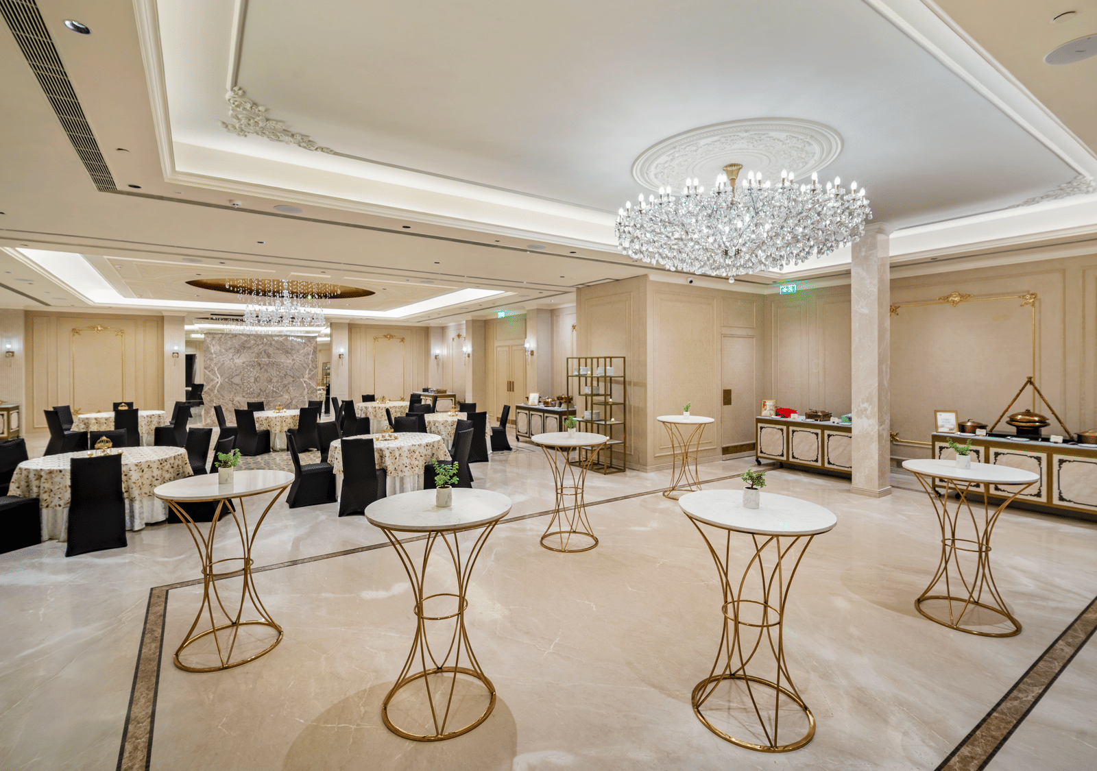 An elegant pre-function or cocktail area at Grand Chennai by GRT Hotels featuring numerous white and black high-top tables, perfect for networking or informal gatherings. A grand, modern chandelier illuminates the spacious room with a soft light.