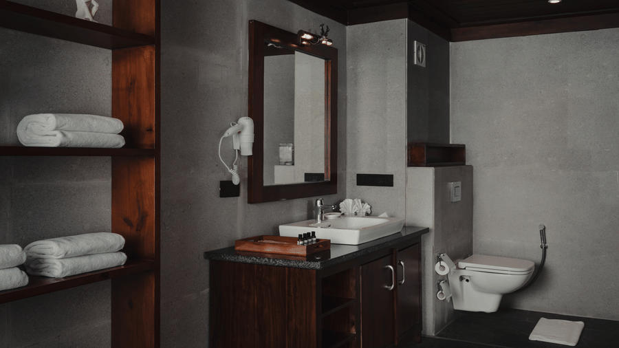 A bathroom at The Ananta Udaipur featuring open storage shelves with stacked towels, vanity unit, washbasin, wall mirror, and toilet.