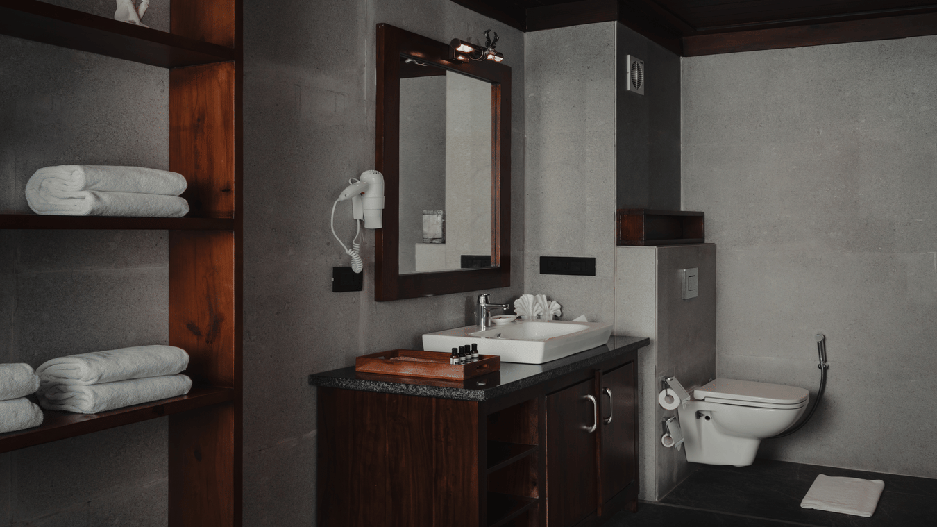 A bathroom at The Ananta Udaipur featuring open storage shelves with stacked towels, vanity unit, washbasin, wall mirror, and toilet.