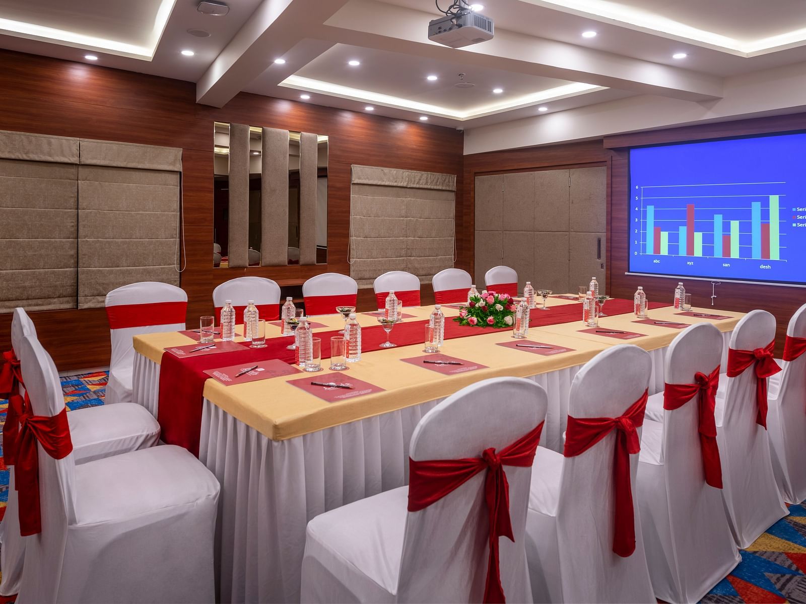 A conference room with a long table surrounded by chairs with red sashes, and a projection screen displaying a presentation.
