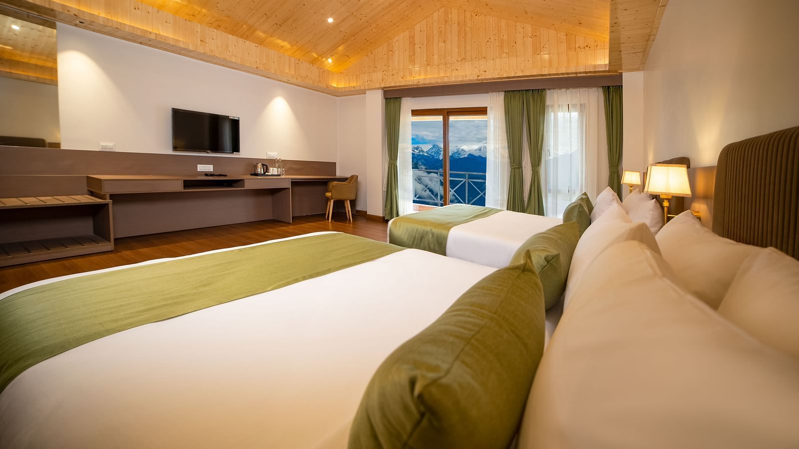 Modern hotel room with two large beds featuring green runners and bolster pillows, wooden flooring and ceiling, a wide work desk with a flat-screen TV, and large windows with curtains revealing a scenic mountain view - Yashshree Pelling Chekhim Retreat.