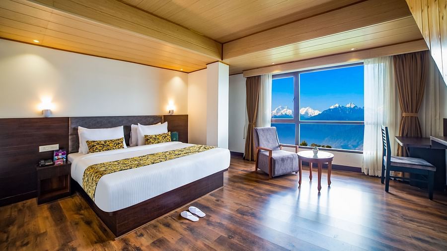 A bright bedroom in Sumitel Hotel with a large window showing an outdoor view, a king-size bed, with a chair and a round table in the corner with wooden flooring around
