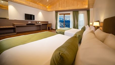 Modern hotel room with two large beds featuring green runners and bolster pillows, wooden flooring and ceiling, a wide work desk with a flat-screen TV, and large windows with curtains revealing a scenic mountain view - Yashshree Pelling Chekhim Retreat.