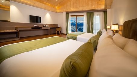 Modern hotel room with two large beds featuring green runners and bolster pillows, wooden flooring and ceiling, a wide work desk with a flat-screen TV, and large windows with curtains revealing a scenic mountain view - Yashshree Pelling Chekhim Retreat.