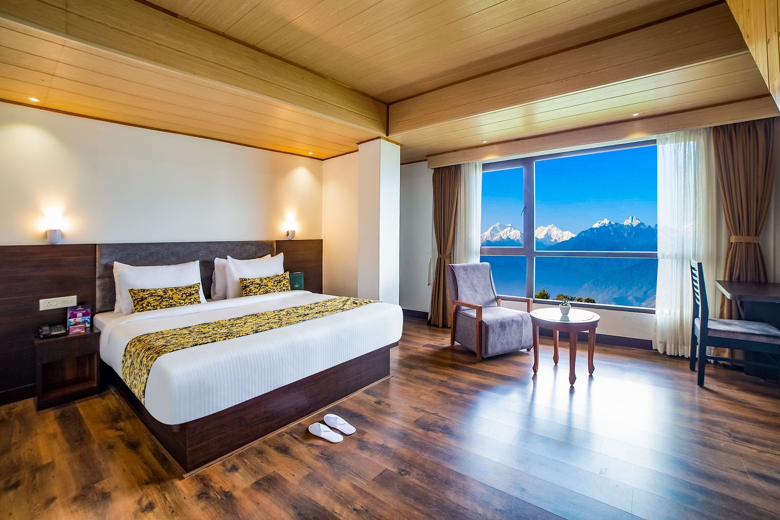 A bright bedroom in Sumitel Hotel with a large window showing an outdoor view, a king-size bed, with a chair and a round table in the corner with wooden flooring around
