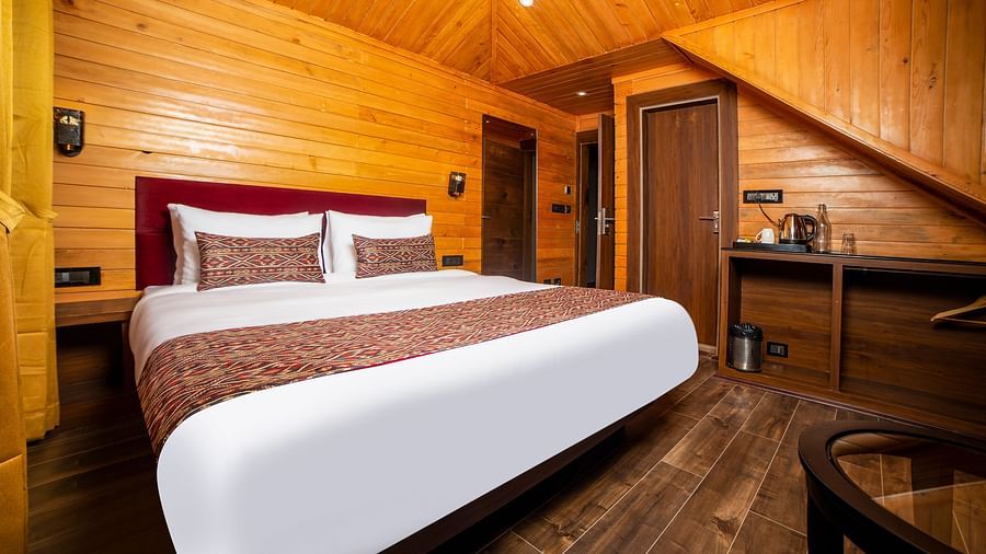 A hotel room with wooden walls, ceiling, and flooring, featuring a large bed with white linens and red-patterned accents, a small kitchenette area with a sink and countertop, and a round glass-top table in the corner  - Yashshree Lachen.