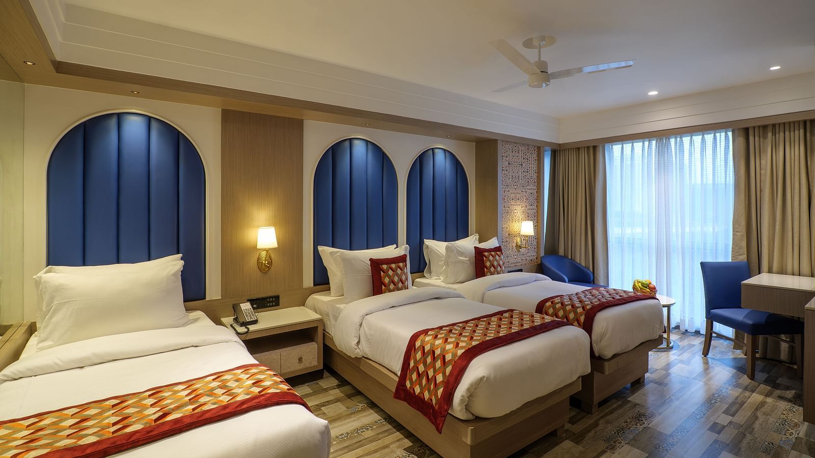 A spacious hotel room including three beds with patterned spreads, blue arched headboards, and a seating area by the window - Melior Inn