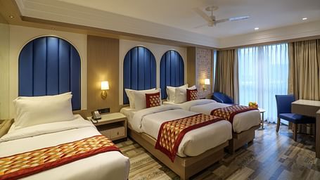 A spacious hotel room including three beds with patterned spreads, blue arched headboards, and a seating area by the window - Melior Inn