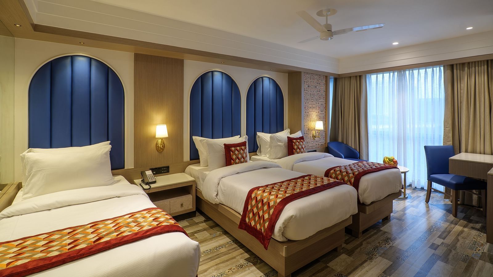 A spacious hotel room including three beds with patterned spreads, blue arched headboards, and a seating area by the window - Melior Inn