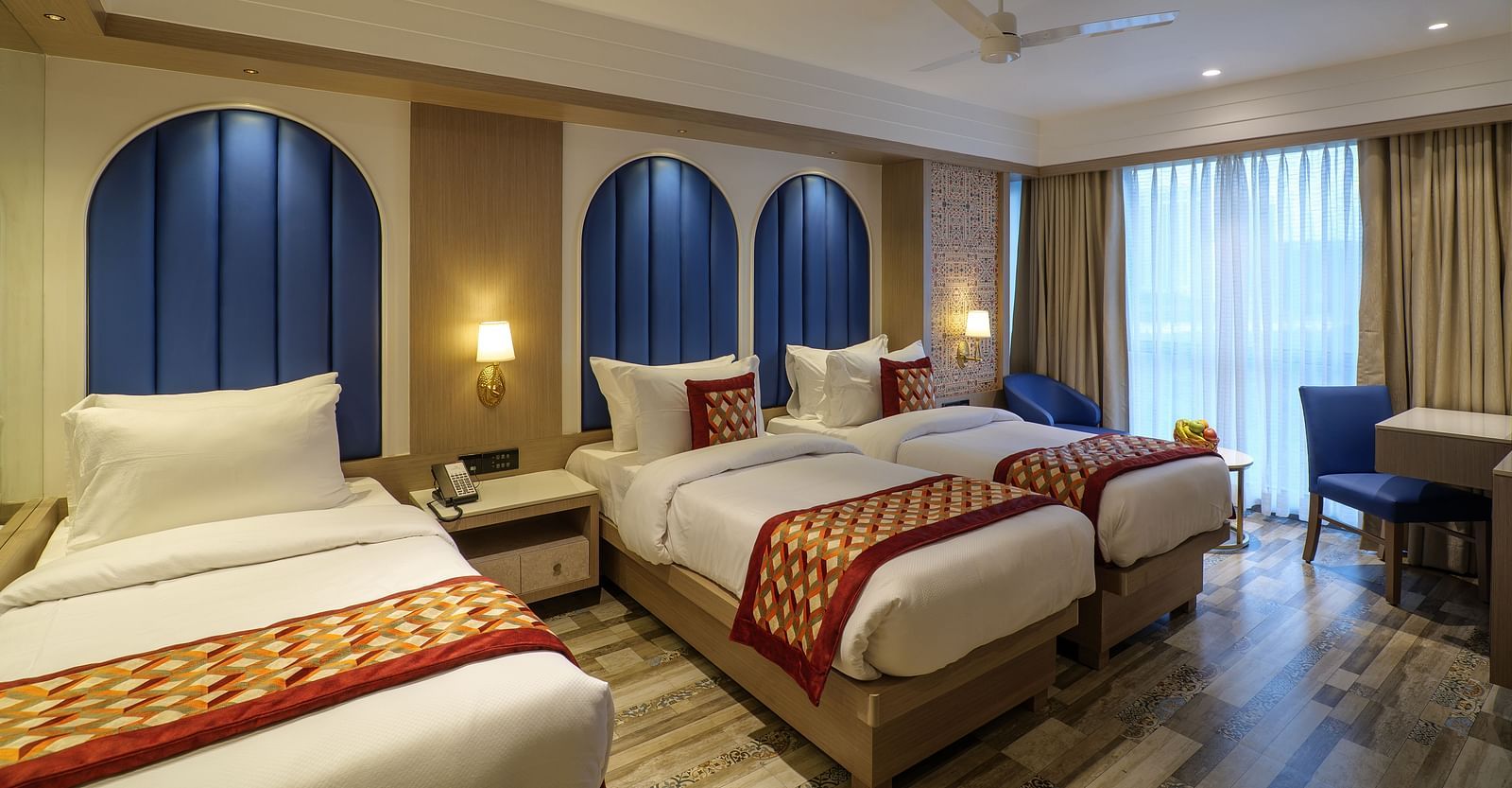 A spacious hotel room including three beds with patterned spreads, blue arched headboards, and a seating area by the window - Melior Inn