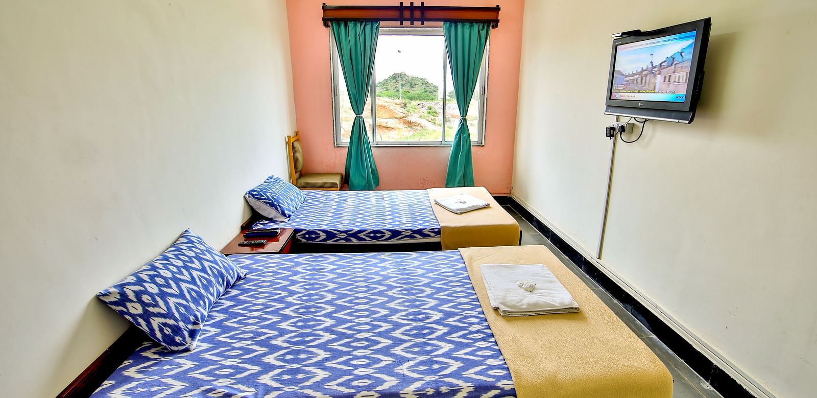 A side view of three beds with bedding in a room with a window and TV at Hotel Sahara - Comfortable Accommodations in Ramoji Film City, Hyderabad.