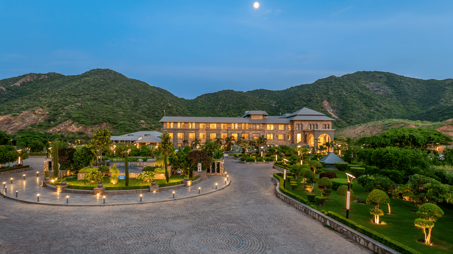 The facade of warmly lit Pushkara Resort & Spa, Ajmer, captured using a drone showcasing its hills in the background and a rising moon.