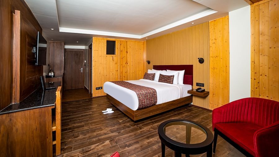 Spacious hotel room with a large bed featuring white and brown bedding, wood-paneled walls and flooring, a wall-mounted TV, a red armchair, and a glass-top coffee table creating a comfortable and modern setting - Yashshree Lachen.