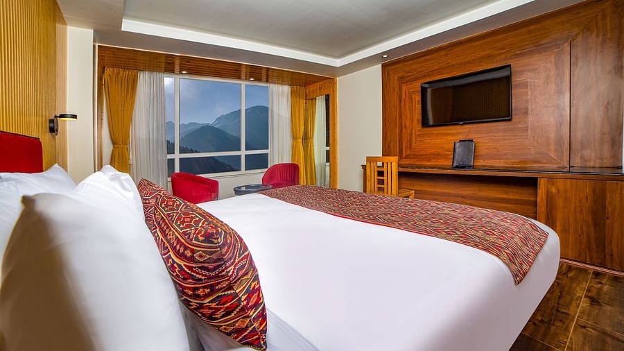 Bright hotel room with a neatly made bed featuring white and patterned bedding, a wooden entertainment unit with a mounted TV, two red chairs by a large window offering a scenic mountain view, and warm wooden decor throughout - Yashshree Lachen.