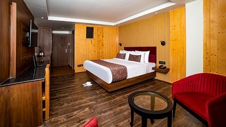 Spacious hotel room with a large bed featuring white and brown bedding, wood-paneled walls and flooring, a wall-mounted TV, a red armchair, and a glass-top coffee table creating a comfortable and modern setting - Yashshree Lachen.