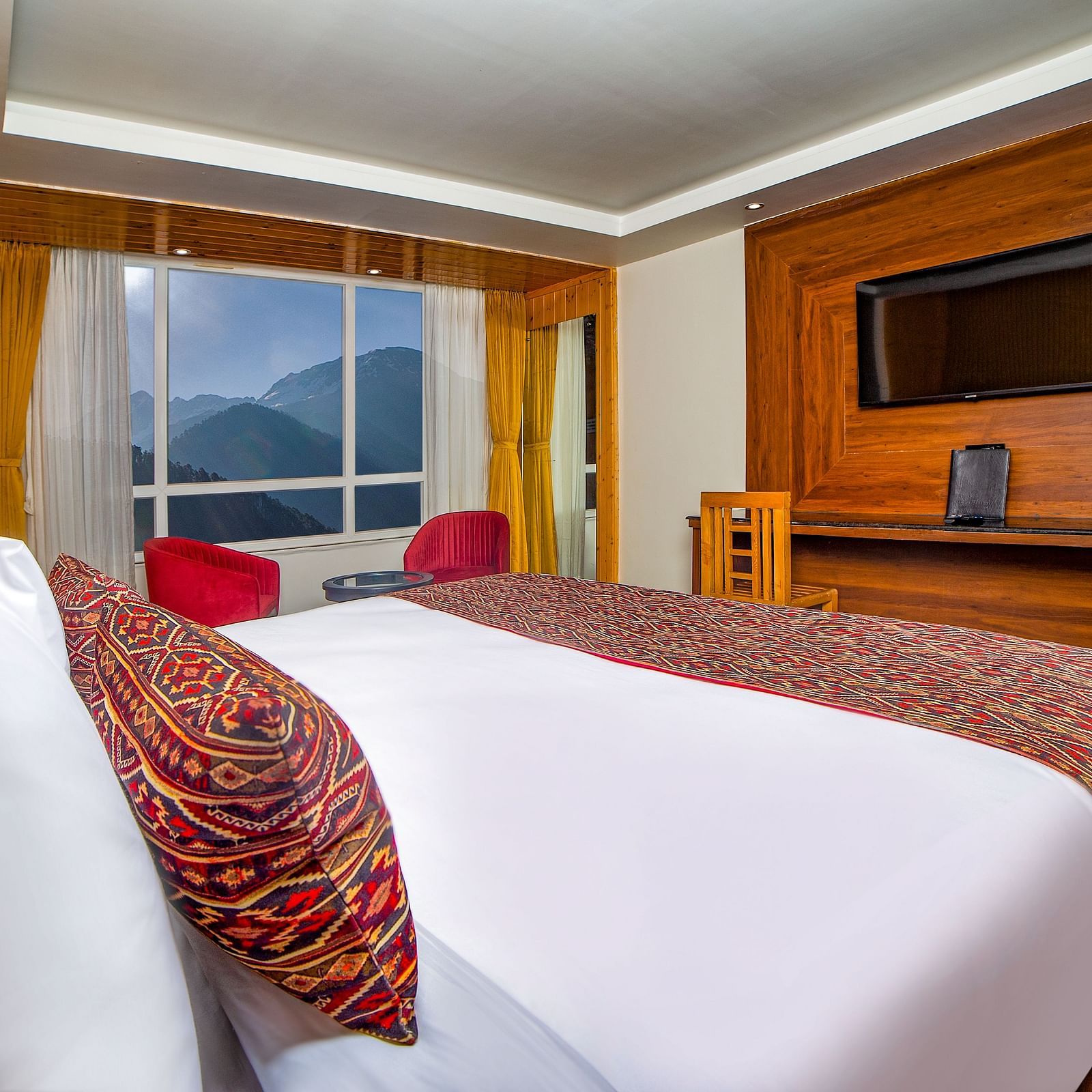 Bright hotel room with a neatly made bed featuring white and patterned bedding, a wooden entertainment unit with a mounted TV, two red chairs by a large window offering a scenic mountain view, and warm wooden decor throughout - Yashshree Lachen.