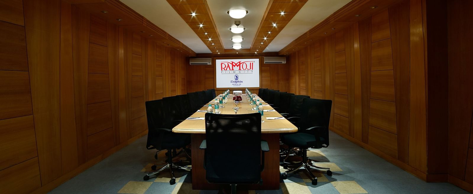 A modern boardroom with a long table, black chairs, and a projector screen at the end. - Tara, Comfort Hotel in Hyderabad - Ramoji Film City.