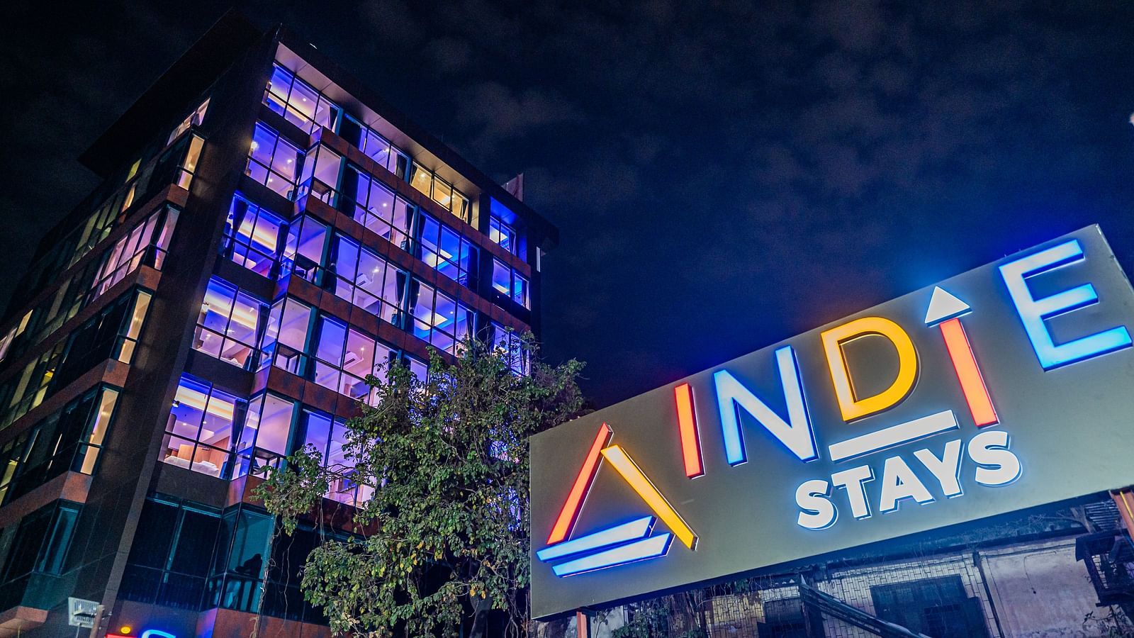 A night view of Indie Stays Mumbai International Airport, showcasing the illuminated name board on one side and the building's striking architecture on the other.