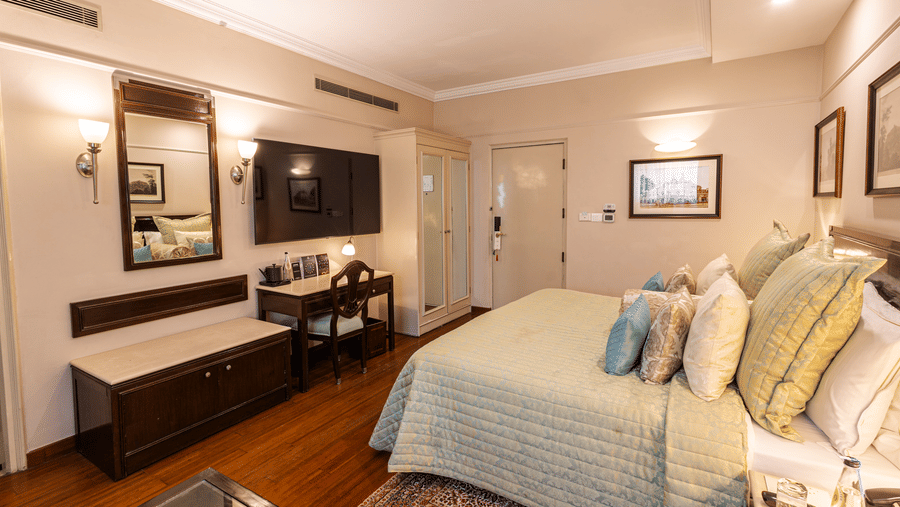 a corner view of the boutique room showcasing the king size bed along with a wall mounted tv and a half rectangular mirror - The Claridges New Delhi