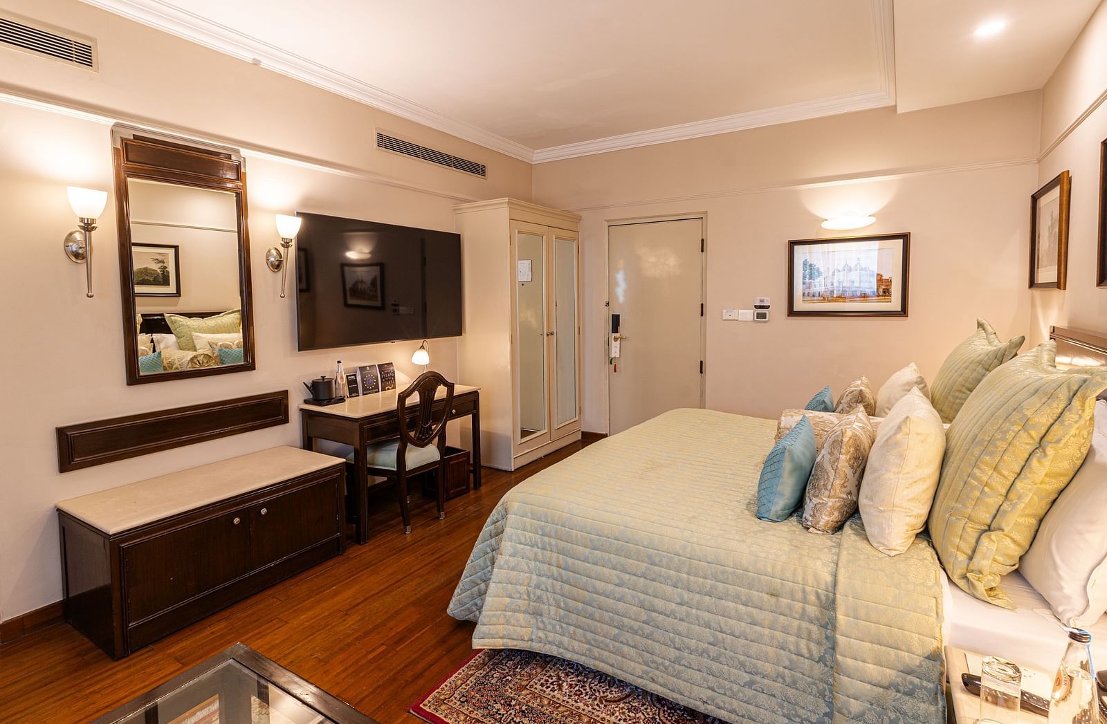 a corner view of the boutique room showcasing the king size bed along with a wall mounted tv and a half rectangular mirror - The Claridges New Delhi