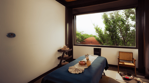 A serene spa treatment room with a massage table ready for use. Natural light streams through a large window offering a view of greenery. A wooden chair sits nearby. The atmosphere is calm and inviting.