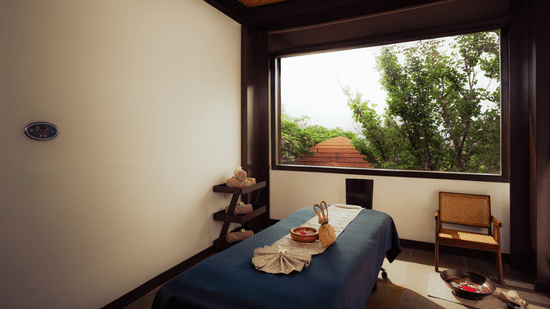 A serene spa treatment room with a massage table ready for use. Natural light streams through a large window offering a view of greenery. A wooden chair sits nearby. The atmosphere is calm and inviting.