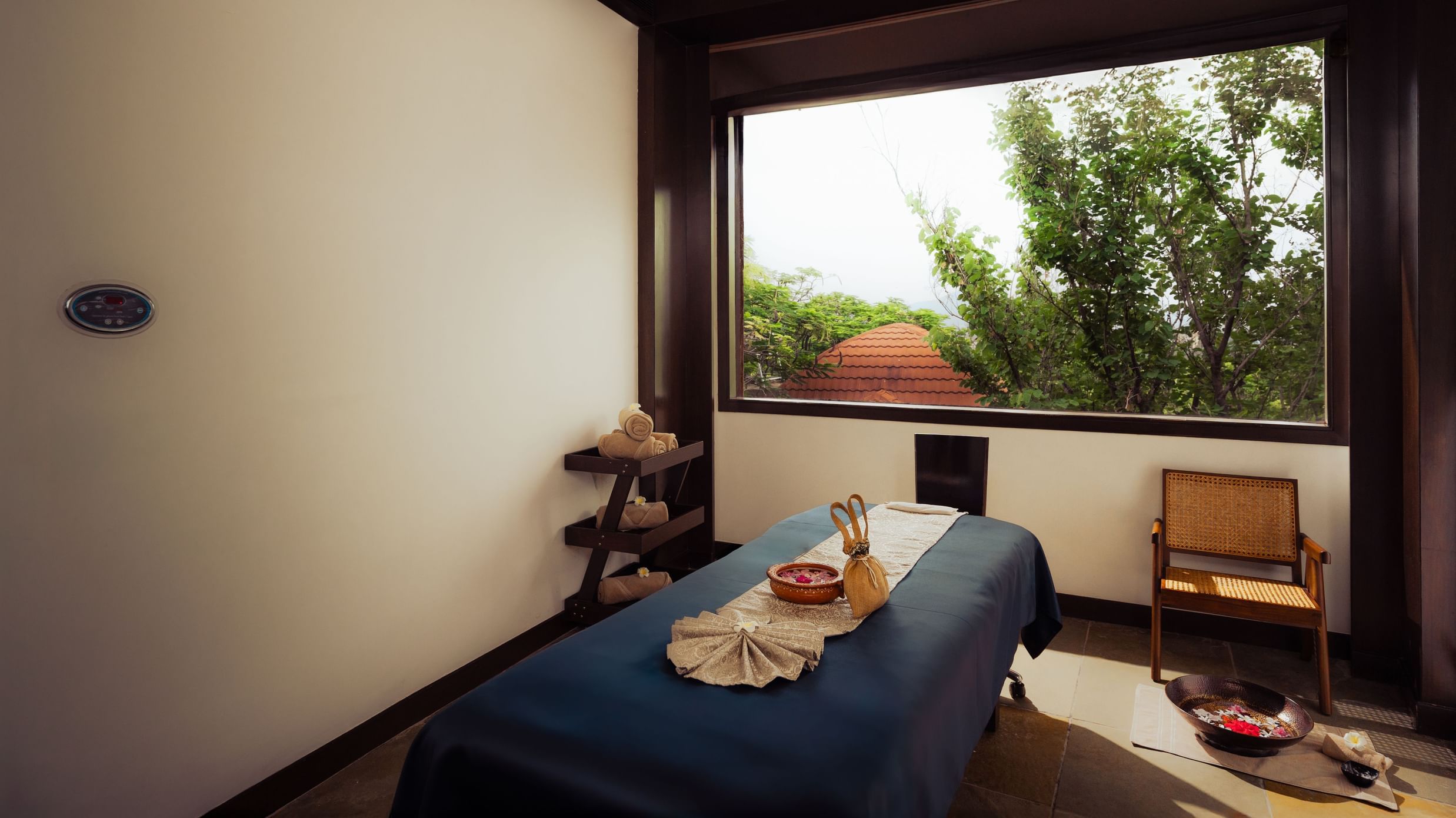 A serene spa treatment room with a massage table ready for use. Natural light streams through a large window offering a view of greenery. A wooden chair sits nearby. The atmosphere is calm and inviting.
