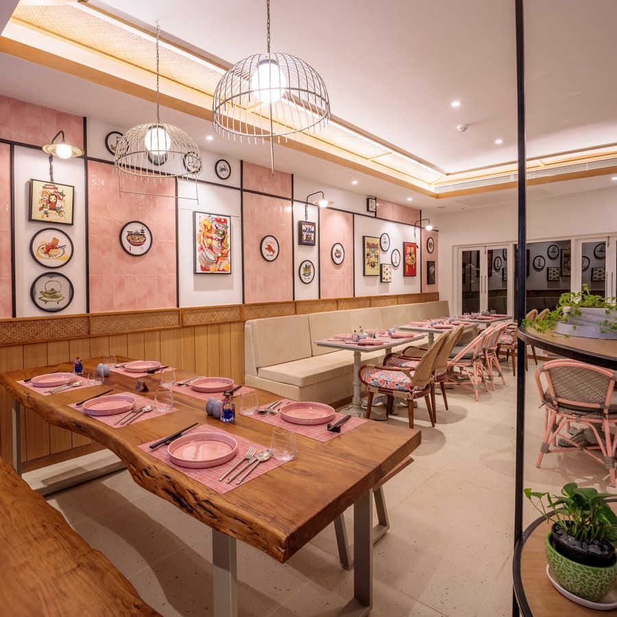A restaurant at Heritage Village Resort and Spa, Goa showcasing wooden table and chairs on one side and on the other there is sofa and a table. Each table has plates, cutlery, and glasses placed. The walls have frames of various dishes.
