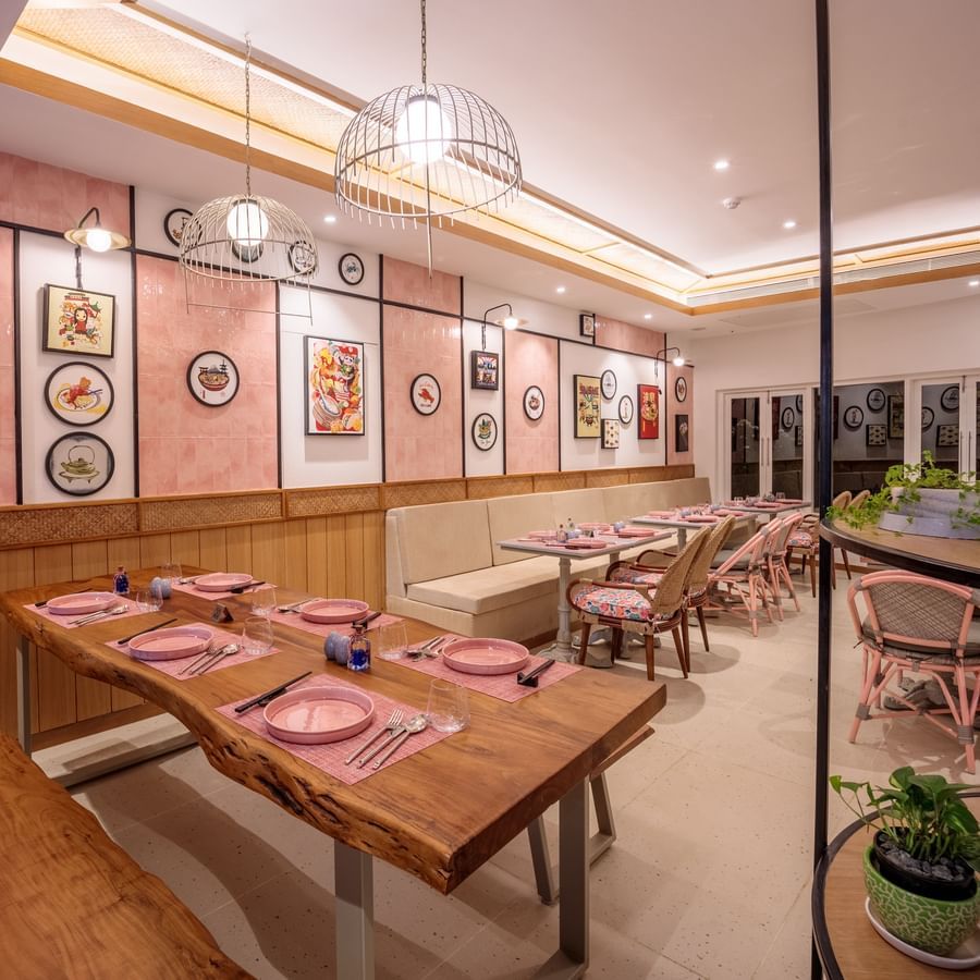 A restaurant at Heritage Village Resort and Spa, Goa showcasing wooden table and chairs on one side and on the other there is sofa and a table. Each table has plates, cutlery, and glasses placed. The walls have frames of various dishes.