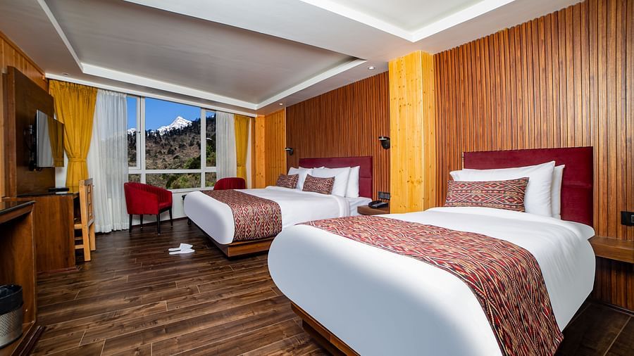 Modern hotel room with two large beds featuring red upholstered headboards and patterned runners, wooden panel walls, hardwood flooring, a flat-screen TV, and large windows showcasing a mountain view - Yashshree Lachen.