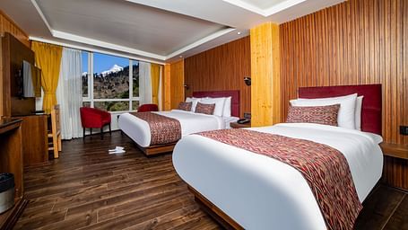 Modern hotel room with two large beds featuring red upholstered headboards and patterned runners, wooden panel walls, hardwood flooring, a flat-screen TV, and large windows showcasing a mountain view - Yashshree Lachen.
