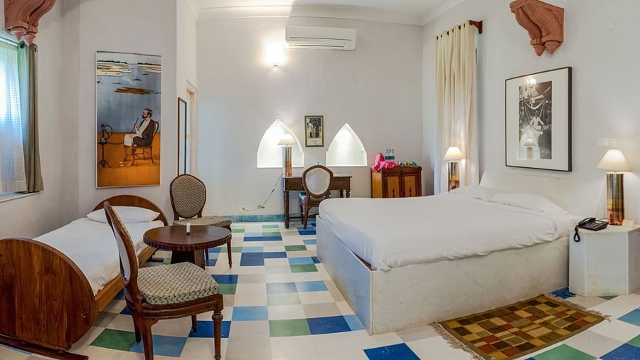 Hotel room with two beds, a desk and chair, a small table with chairs, patterned flooring, and artwork on the walls at Tijara Fort-Palace, Alwar.