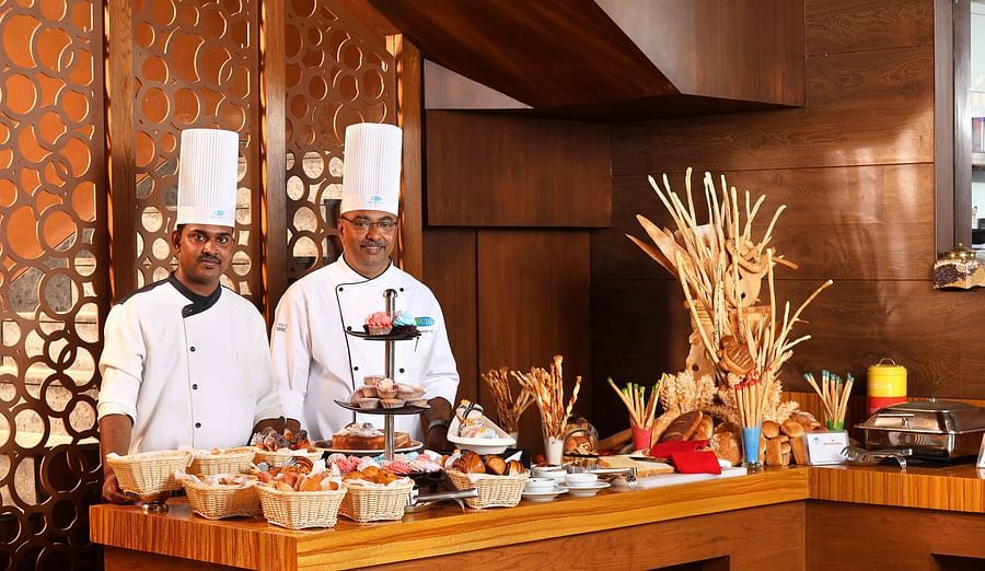 Two men in white chef uniforms and tall white hats stand behind a buffet table filled with various pastries, breads, and desserts - UDS Group of Hotels.