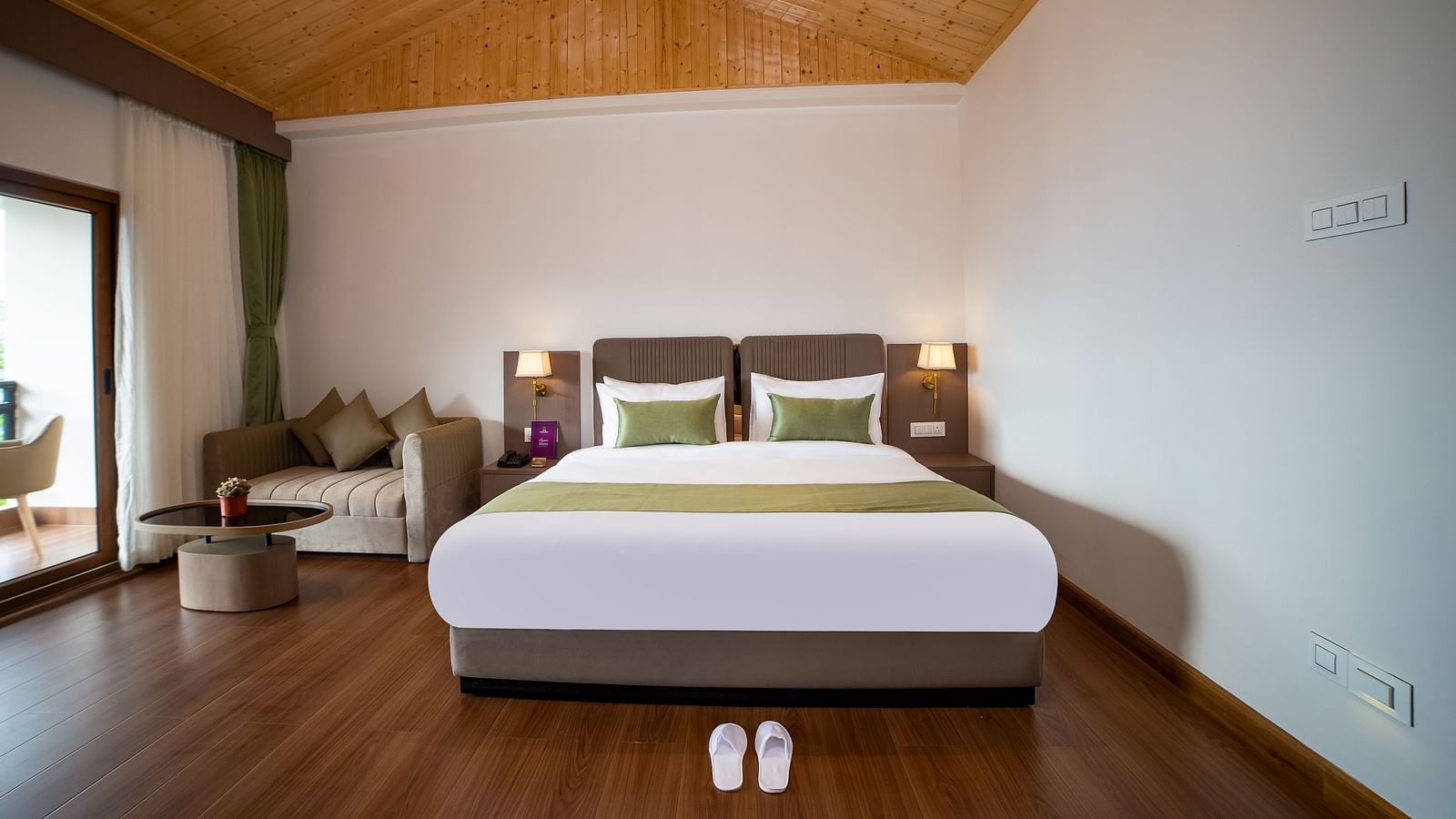 Modern hotel room featuring a large bed with green accents, wooden flooring, vaulted wooden ceiling, a cosy seating area, and a balcony door allowing natural light - Yashshree Pelling Chekhim Retreat.