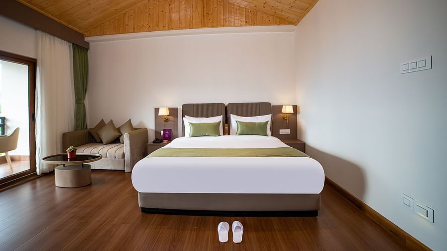 Modern hotel room featuring a large bed with green accents, wooden flooring, vaulted wooden ceiling, a cosy seating area, and a balcony door allowing natural light - Yashshree Pelling Chekhim Retreat.