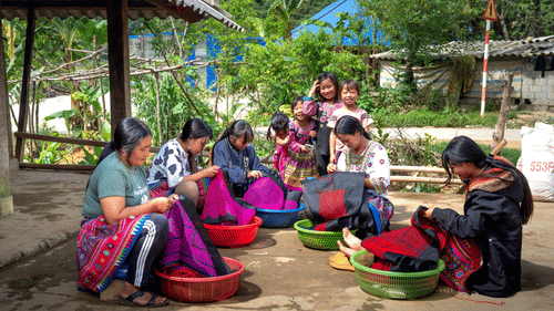 a group of women dyeing sarees in a plastic bowl that is filled with dye while the children look on