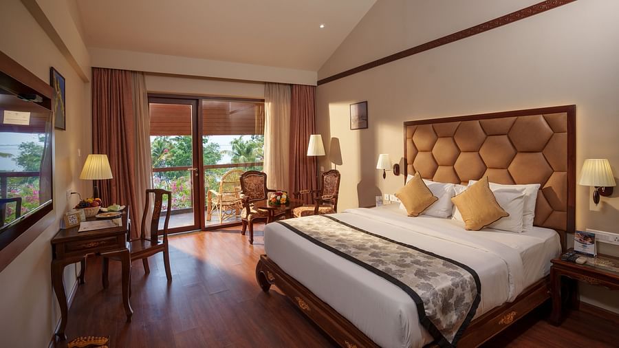 Lake View Elite Room at Uday Backwater Resort featuring a plush bed in front of the TV mounted on the wall and a work desk facing the wall. A sitting space has been created next to the door to the balcony.