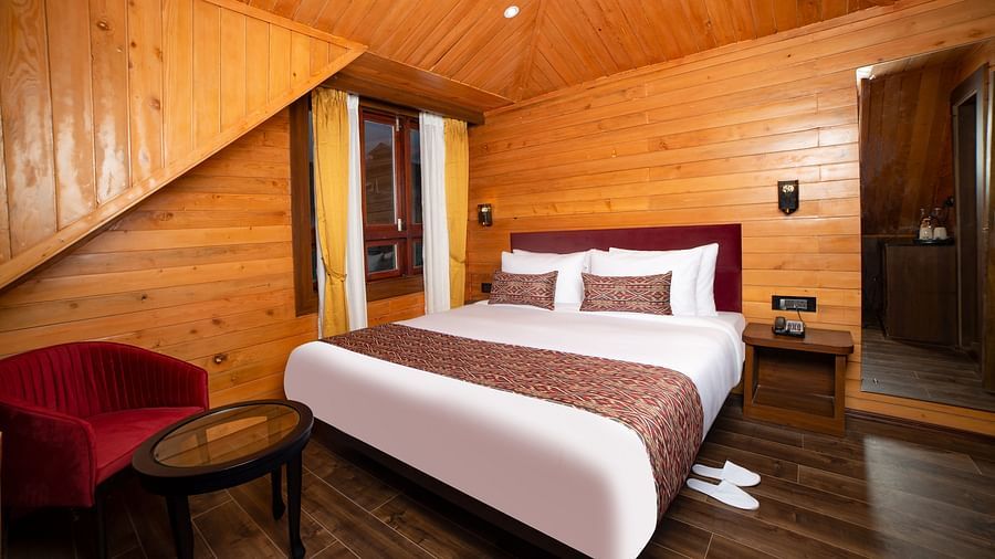A hotel room with wooden walls and sloped ceiling, featuring a large bed with white linens and red accents, a red armchair with a round glass-top table, and a wooden nightstand with a wall-mounted lamp - Yashshree Lachen.
