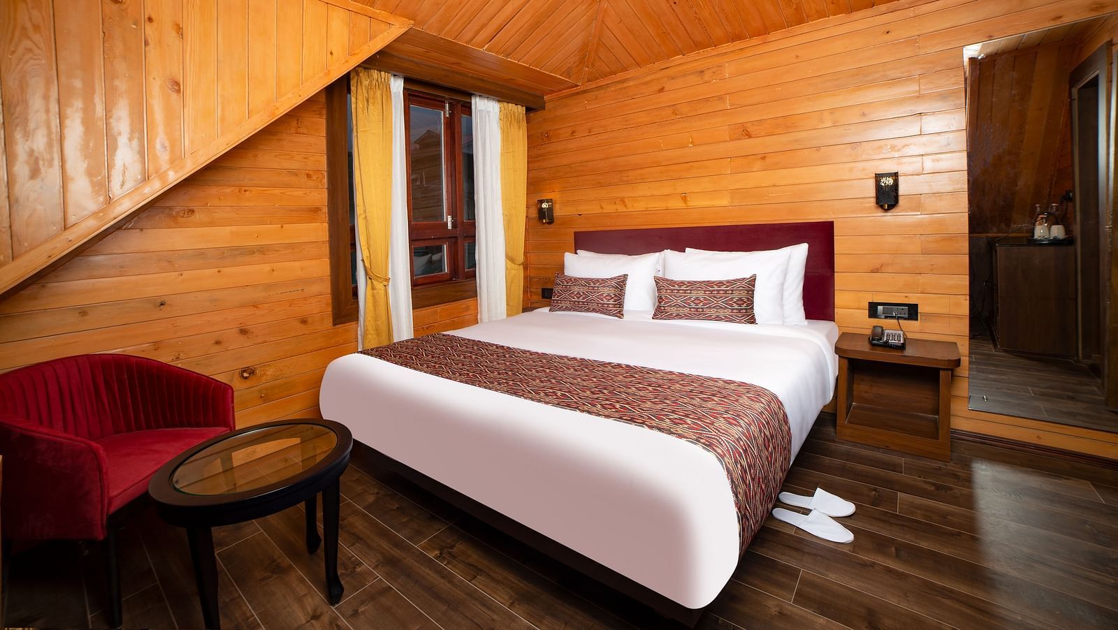 A hotel room with wooden walls and sloped ceiling, featuring a large bed with white linens and red accents, a red armchair with a round glass-top table, and a wooden nightstand with a wall-mounted lamp - Yashshree Lachen.